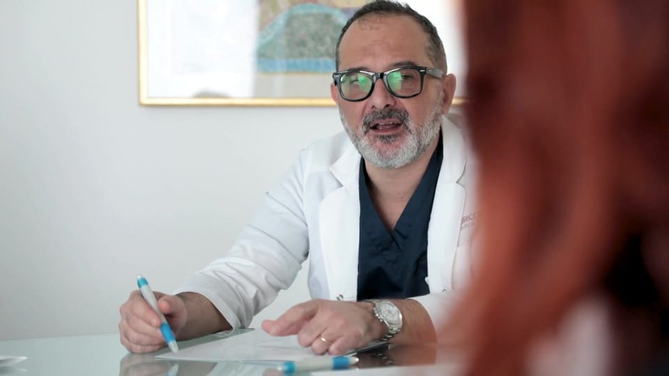 Il Prof. Coricciati alla scrivania, in camice, mentre parla con una paziente di spalle e scrive