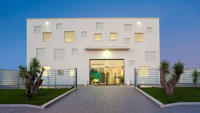 La sede di Martano all’ora blu, di fronte: il cubo bianco a finestre quadrate, l’ingresso illuminato, due ulivi secolari ai lati del viale